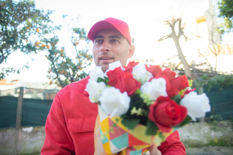 A man in a red cap and matching red jacket holding a floral bouquet with red and white roses, wrapped in colorful paper. The bouquet is positioned in front of him, slightly obscuring his waist. He is