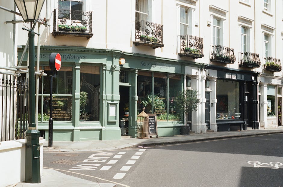 A floral shop with a light green exterior and large glass windows displaying an assortment of fresh flowers and plants inside. The shop has decorative black wrought-iron balconies above with small pot