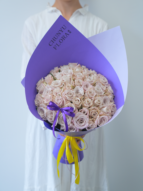 A person dressed in a white shirt holds a large floral bouquet wrapped in vibrant purple and yellow paper. The bouquet features numerous pale pink and cream roses, densely arranged to create a rounded