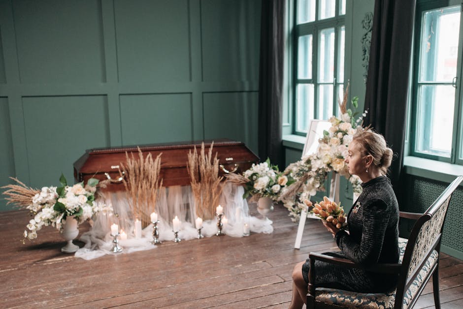 A woman dressed in a black textured outfit is sitting on an ornate wooden chair in a softly lit room with green paneled walls and large windows. She is holding a floral bouquet featuring peach-colored