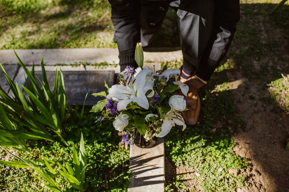 A floral arrangement featuring white lilies, purple and white tulips, and small purple and white filler flowers, displayed in a dark glass vase. The bouquet is held by a person wearing dark clothing a
