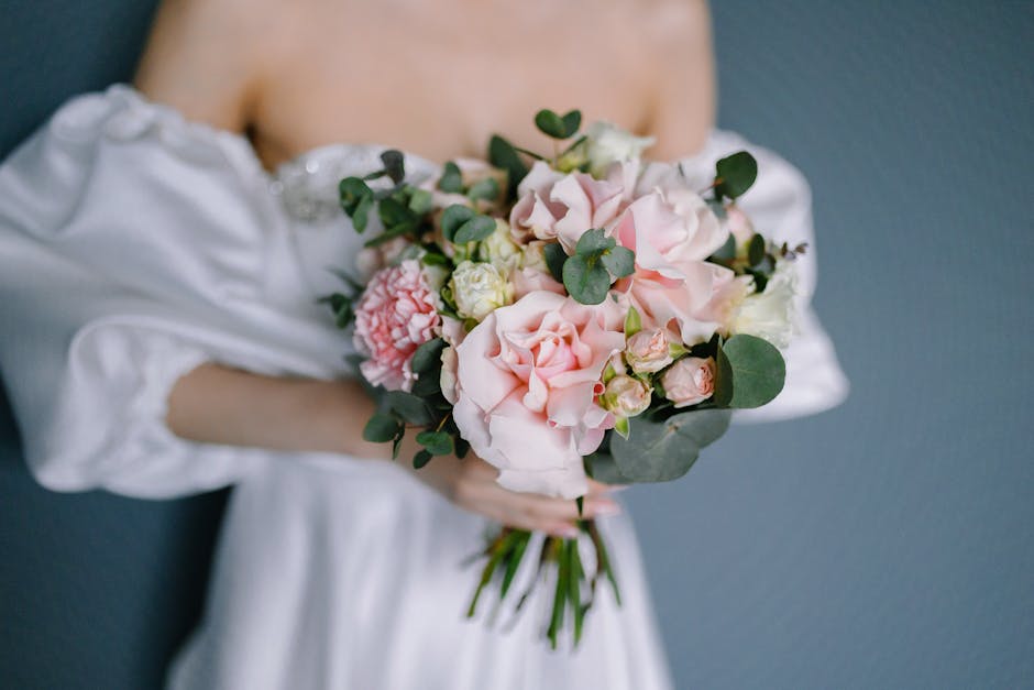 A person in a white off-the-shoulder dress holds a bouquet of fresh flowers, featuring large pale pink roses, white lisianthus, and smaller pink carnations, accented with green eucalyptus leaves. The 