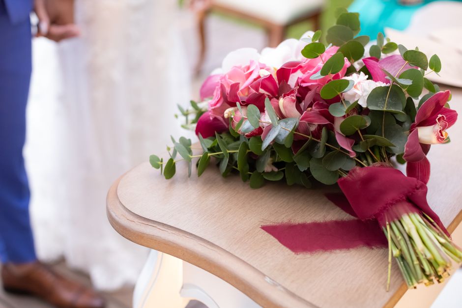 A bouquet of fresh pink and white orchids accented with green eucalyptus leaves is arranged on a light wooden table, wrapped with burgundy ribbon. The flowers are vibrant and healthy with smooth petal